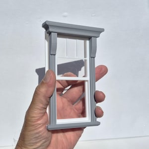 May include: A miniature window frame featuring a grey exterior casing with decorative molding at the top and bottom. The interior section is white, showcasing a classic double-hung style design with a horizontal divider bar across the center.
