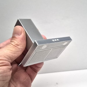 May include: A miniature silver range hood with a rectangular design. The hood features two square filters and three small circular lights. The object is held in a hand.