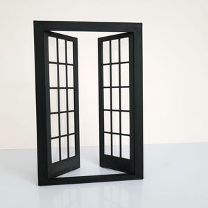 May include: A black, miniature window frame with two open doors. Each door has a grid of small, rectangular panes. The frame is rectangular and sits upright on a white surface, against a white background. The doors are slightly ajar.