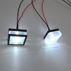 May include: Two illuminated, square-shaped LED lights with black frames and white, glowing interiors. Each light is connected to wires with red and black insulation.