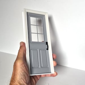 Pode incluir: Uma porta cinzenta em miniatura com moldura branca, apresentando uma janela de seis painéis no topo e dois painéis retangulares na parte inferior. A porta inclui um pequeno puxador preto no lado direito, concebida para casas de bonecas.