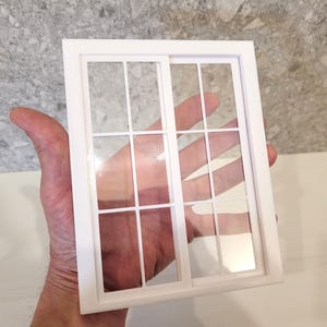 Op de afbeelding: Een wit miniatuurraam met een rasterpatroon, vastgehouden in een hand. Het raam heeft een rechthoekig frame en heldere ruiten, wat duidt op een poppenhuis of model. Het ontwerp bevat een schuifraamelement.