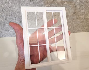 May include: A white miniature window with a grid pattern, held in a hand. The window has a rectangular frame and clear panes, suggesting a dollhouse or model. The design includes a sliding window element.