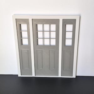 Pode incluir: Uma porta cinza em miniatura com moldura branca. A porta tem um painel central com uma grade de pequenos vidros e dois painéis laterais com janelas retangulares. O design é simples e simétrico, adequado para uma casa de bonecas ou um projeto de artesanato.