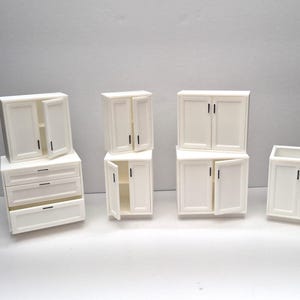 Puede incluir: Colección de armarios y cajones blancos en miniatura. Algunos armarios tienen puertas abiertas, revelando el interior. La ferretería es negra. El conjunto incluye una cómoda y varios estilos de armarios.