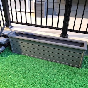 May include: A rectangular, gray planter box with a dark interior sits on a green surface. The box has horizontal lines and a light gray trim. A black railing and steps are in the background.