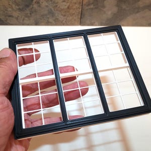 Op de afbeelding: Een zwart-wit miniatuur raamkozijn met een rasterpatroon. Het frame wordt door een hand vastgehouden, waarbij het gedetailleerde ontwerp wordt getoond. Het raam is verdeeld in meerdere ruiten, geschikt voor poppenhuizen of modelbouwprojecten.