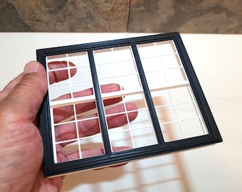 May include: A rectangular miniature window frame featuring a black outer border and a white interior grid pattern. The frame is divided into three vertical sections, each containing a grid of smaller square panes, creating a classic architectural look.