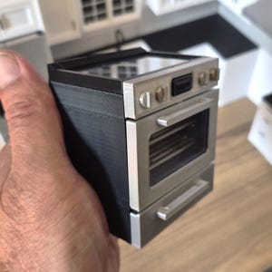 Könnte beinhalten: Ein Miniatur-Küchenherdmodell mit schwarzem Rahmen, einer silberfarbenen Frontplatte mit verglaster Ofentür, einer darunter liegenden Schublade und einer schwarzen Kochfeldoberfläche mit silbernen Drehknöpfen an der Oberkante.