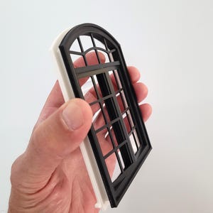 May include: A small black and white window with an arched top and a grid pattern. The window is held in a hand, highlighting its intricate design. The frame is white, and the window panes are black.