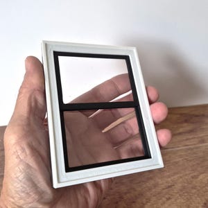 Può includere: Un piccolo telaio di finestra rettangolare con bordo bianco e interno nero. La finestra è divisa in due vetri da una barra nera orizzontale. Il telaio è tenuto in mano su uno sfondo bianco.