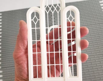 May include: A white, arched window frame with three sections, held in a hand. The frame has a detailed lattice design and is made of a solid, opaque material. The background shows a gray, brick-like surface.