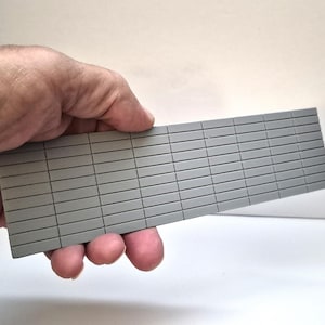 May include: A rectangular grey plastic sheet featuring a grid pattern of small, uniform, horizontal bricks. The surface is flat and matte, with recessed lines creating the appearance of a miniature brick wall texture, suitable for scale modelling projects.