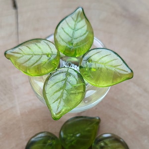 May include: A set of seven green glass leaf-shaped beads. The beads are translucent and have a delicate vein pattern.