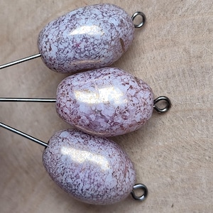 Könnte beinhalten: Drei ovale Perlen mit gesprenkeltem, perlmuttartigem Finish in Lavendel- und Weißtönen. Jede Perle hat eine silberfarbene Metallschlaufe und einen Draht, der sich von der Basis erstreckt. Die Perlen sind auf einer hellbraunen Oberfläche angeordnet.