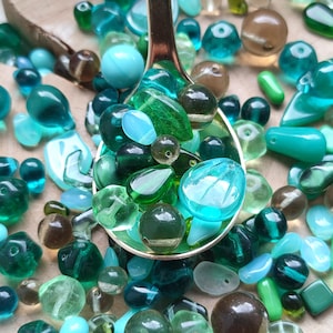 Peut inclure: Un assortiment de perles de verre dans des tons de turquoise, vert et marron, disposées sur une surface en bois. Une cuillère dorée contient une sélection de perles, mettant en valeur leurs différentes formes et tailles.
