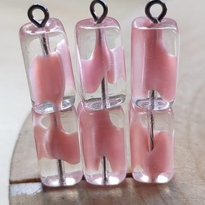 Op de afbeelding: Zes heldere, rechthoekige kralen met een roze, abstract ontwerp aan de binnenkant. Elke kraal heeft een zilverkleurige draad die erdoorheen loopt, bekroond met een zwarte lus. De kralen zijn gestapeld op een houten oppervlak.