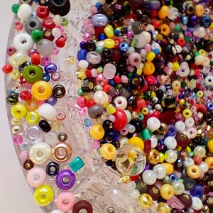 May include: A close-up of a clear glass bowl filled with a variety of colorful beads. The beads are in different shapes and sizes, including round, square, and oval. The beads are arranged in a random pattern, creating a visually appealing and textured surface.