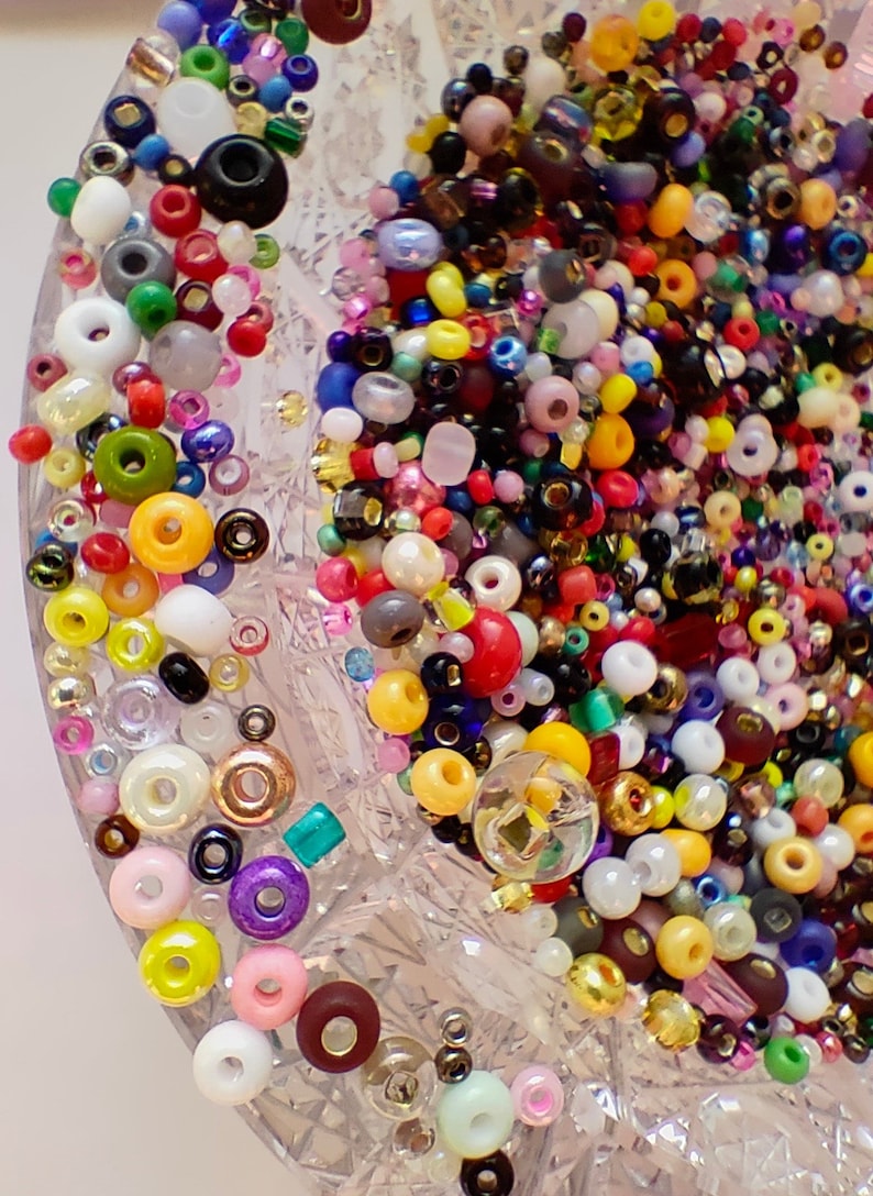 May include: A close-up of a clear glass bowl filled with a variety of colorful beads. The beads are in different shapes and sizes, including round, square, and oval. The beads are arranged in a random pattern, creating a visually appealing and textured surface.