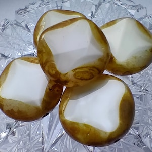 May include: Four white and gold glass beads with a square shape and rounded edges. The beads have a white center and a gold rim.