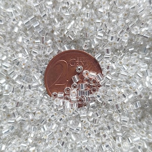 Può includere: Una grande collezione di piccole perline di vetro cilindriche e trasparenti con un interno argentato. Una moneta da 2 euro color rame è posizionata tra le perline per fornire una scala delle dimensioni di questi componenti per il fai da te.
