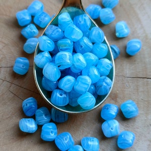 Könnte beinhalten: Ein Löffel gefüllt mit hellblauen und weißen, marmorierten Glasperlen. Die Perlen sind um den Löffel herum auf einer Holzoberfläche verstreut.