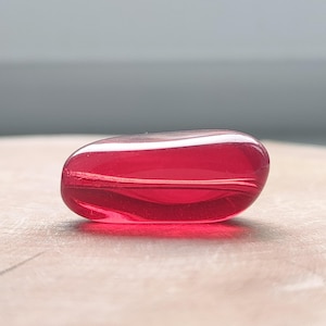Könnte beinhalten: Eine glatte, durchscheinende, tiefrote Glasperle in einer länglichen, kieselsteinartigen Form. Die Perle hat ein horizontal verlaufendes Loch in der Mitte, wobei sich das Licht auf ihrer polierten, gewölbten Oberfläche bricht und die Farbe betont.