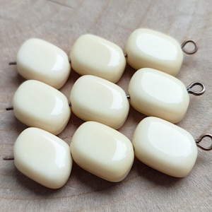May include: Nine off-white, oval-shaped beads with a glossy finish. Each bead has a small, dark metal loop or pin attached. The beads are arranged on a light brown wooden surface, suggesting a craft or jewelry-making theme.