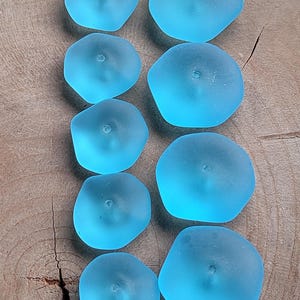 May include: Ten light blue, frosted glass beads arranged in two vertical columns. Each bead has an irregular, rounded shape with a smooth, matte texture and a single, centred hole for threading, displaying a translucent, sea-glass appearance.