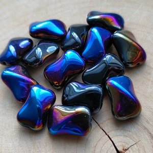 Op de afbeelding: Een verzameling van iriserende glazen kralen in tinten blauw, zwart en paars. De kralen zijn traanvormig en hebben een gladde, glanzende afwerking.