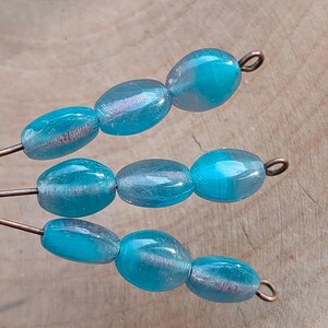 May include: A strand of turquoise blue glass beads with silver flecks. The beads are oval shaped and have a slightly iridescent finish. The beads are strung on a wire with a copper wire loop at each end.