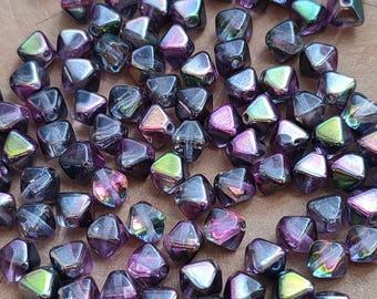 May include: A collection of iridescent, faceted beads in shades of purple, pink, and green. The beads have a geometric shape with multiple sides and a hole for stringing. They are scattered on a wooden surface.