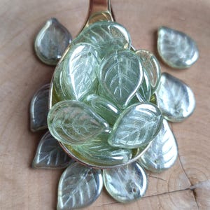 May include: A gold spoon filled with translucent green glass leaf-shaped beads. The beads have a subtle shimmer and are perfect for jewelry making or crafting.