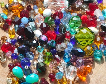 May include: A collection of colorful beads in various shapes and sizes. The beads are in shades of red, green, blue, yellow, and clear, with faceted and smooth surfaces. Some beads have an iridescent sheen, and others are opaque.
