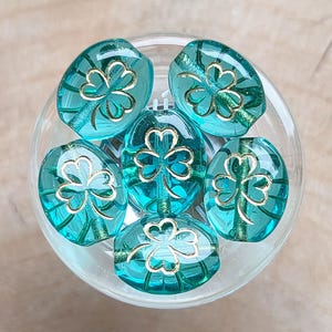 May include: Six teal blue glass beads with gold shamrock designs. The beads are in a clear glass bowl.