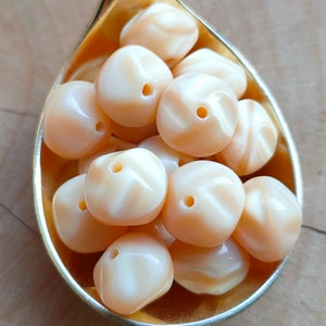 May include: A close-up of a gold spoon filled with peach-colored, faceted, round beads. The beads have a white swirl pattern.