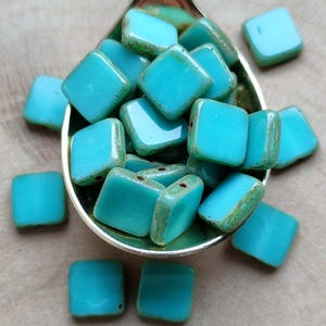 Può includere: Un cucchiaio pieno di perline quadrate di vetro blu turchese. Le perline hanno una superficie leggermente strutturata e sono sparse intorno al cucchiaio.