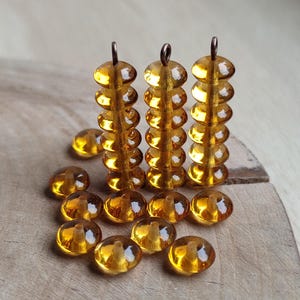 May include: Amber-coloured glass beads with a copper-coloured loop at the top. The beads are arranged in three vertical stacks and scattered on a wooden surface. The beads have a glossy finish and a rounded shape, ideal for jewellery making.