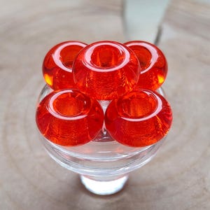 Könnte beinhalten: Fünf durchscheinende, rote Glasperlen, die in einer Gruppe angeordnet sind. Jede Perle hat eine hohle Mitte und sitzt auf einem Sockel aus klarem Glas. Die Perlen sind ein leuchtendes, glänzendes Rot, der Hintergrund ist hellbraun.