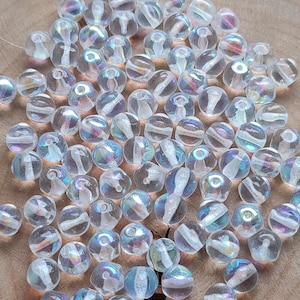 May include: A collection of clear, iridescent glass beads with a swirling, internal design. The beads are spherical and vary slightly in size, scattered on a wooden surface. The beads reflect light, displaying a range of colors.