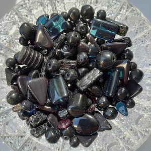 Può includere: Una collezione di perline di vetro nero di varie forme e dimensioni, tra cui design rotondi, rettangolari e sfaccettati. Alcune perline hanno un tocco di blu o viola. Le perline sono esposte in una ciotola di cristallo trasparente.