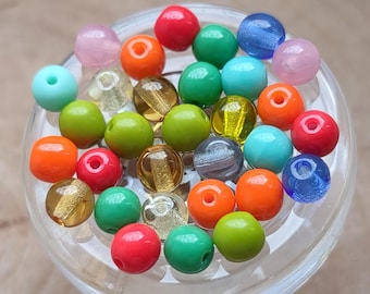 May include: A collection of colorful glass beads in various shapes and sizes, including round, oval, and teardrop. The beads are arranged in a glass bowl and feature a variety of colors, such as red, green, blue, yellow, and pink.