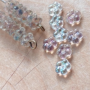 Peut inclure: Gros plan sur des perles transparentes irisées en forme de fleurs. Deux brins de perles sont enfilés sur du fil d'argent avec des boucles dorées. Des perles en vrac sont éparpillées sur une surface en bois, présentant une gamme de couleurs.
