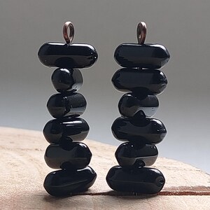 May include: A pair of black beaded earrings. Each earring has a stack of glossy, oval-shaped black beads, connected by a copper-coloured wire loop at the top. The beads are arranged in a vertical column, creating a minimalist, elegant design.