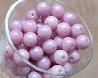 May include: A collection of round, light purple beads with a glossy finish, displayed inside a clear glass container. The beads have a speckled appearance and are evenly sized. The glass container is partially visible, showing its curved shape.