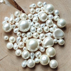 Puede incluir: Una colección de perlas de color marfil de varios tamaños y formas, incluyendo redondas, en forma de lágrima e irregulares. Las perlas están dispersas sobre una superficie de madera natural, creando un contraste de texturas y tonos. Las perlas varían en tamaño, con un brillo lustroso.