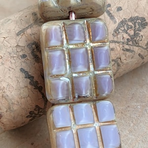 Pode incluir: Três contas quadradas de vidro com um padrão de grade. As contas são de cor púrpura clara com um brilho metálico dourado.