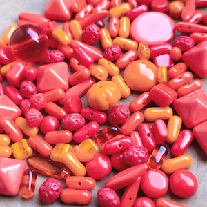 Könnte beinhalten: Eine Sammlung von roten, orangefarbenen und rosa Perlen in verschiedenen Formen. Die Auswahl umfasst runde, längliche, facettierte und geschnitzte Perlen, ideal für Schmuckherstellung oder Bastelprojekte.