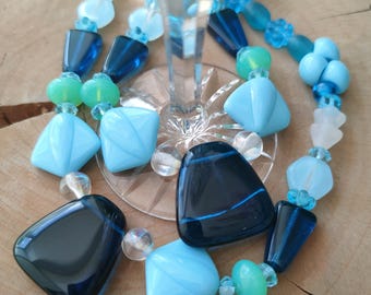May include: A necklace made of blue, green, and white glass beads. The beads are various shapes, including squares, triangles, and ovals. The necklace is draped over a glass wine glass.