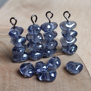 Peut inclure: Gros plan sur plusieurs piles de perles translucides bleu-gris en forme de cœur avec des boucles en métal foncé. Quelques perles en vrac sont dispersées à proximité. Les perles ont une finition brillante et sont disposées sur une surface en bois, idéales pour la création de bijoux.